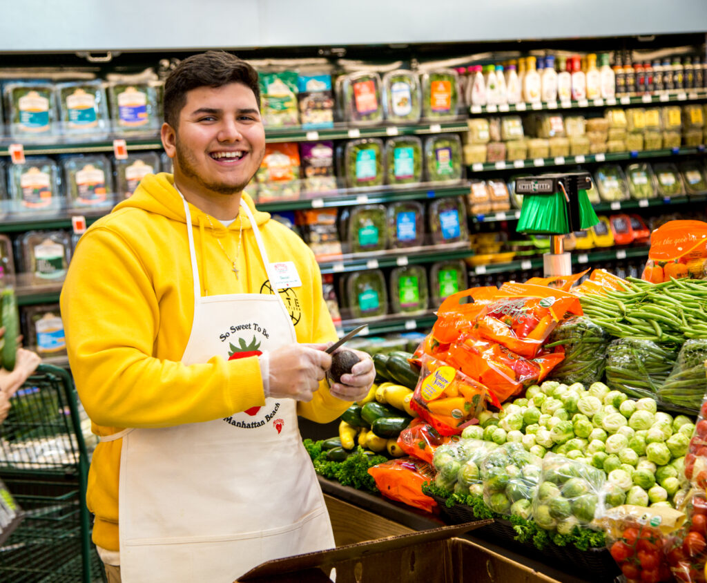 smiling young man wearing apron in produce section of mothers supermarket